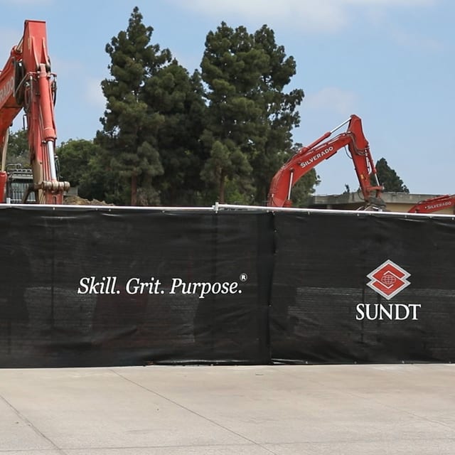 Contractor-branded fence screen on a jobsite fence with excavators behind it